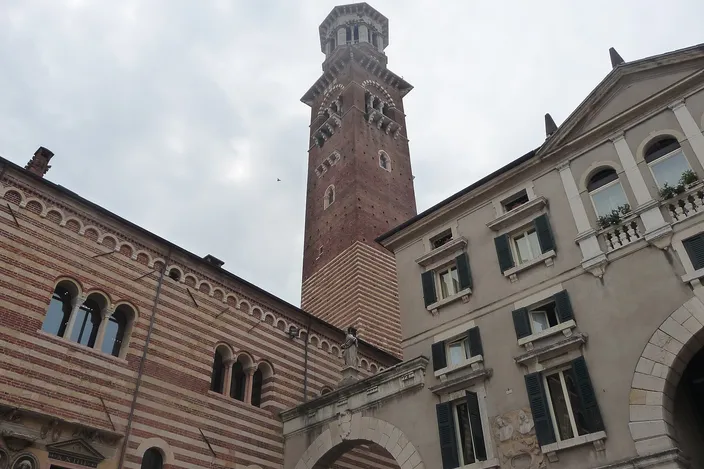 A daytime view in Piazza dei Signori looking up at the tall brick Torre dei Lamberti, flanked by the striped medieval facade of the Palazzo della Ragione on the left and a Renaissance building on the right.