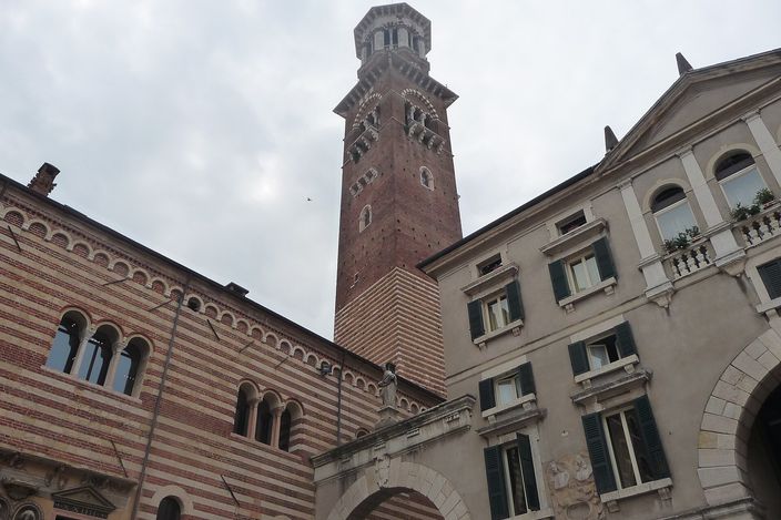 A daytime view in Piazza dei Signori looking up at the tall brick Torre dei Lamberti, flanked by the striped medieval facade of the Palazzo della Ragione on the left and a Renaissance building on the right.