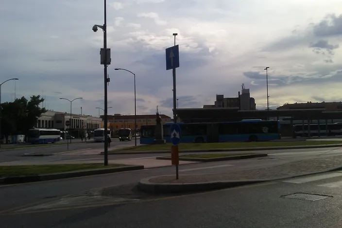 A city bus terminal near Verona's center with several ATV buses boarding passengers, a cyclist and a pedestrian crossing the plaza under an evening sky.
