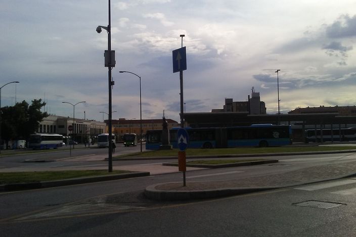 A city bus terminal near Verona's centre with several ATV buses boarding passengers, a cyclist and a pedestrian crossing the plaza under an evening sky.