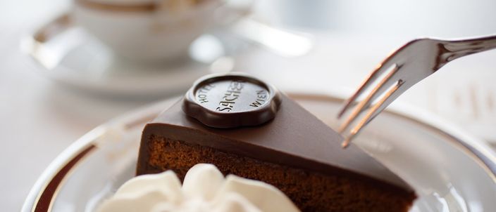 A slice of Sachertorte carrying the round Original Sacher-Torte chocolate seal, a swirl of unsweetened whipped cream beside it on a gold-rimmed plate, a fork entering the glaze and a melange blurred behind.