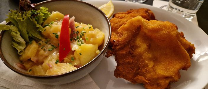 A golden breaded Wiener Schnitzel on a white plate beside a bowl of Viennese potato salad with green leaves