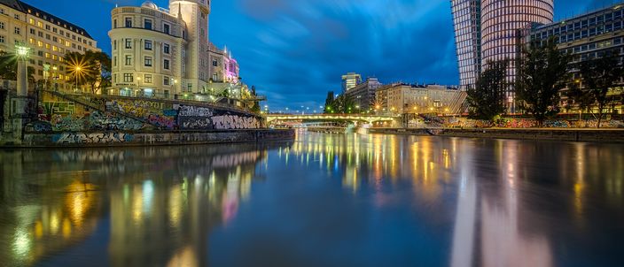 Blue-hour view along the Donaukanal in central Vienna: the ornate Urania observatory building with its little dome on the left, colourful reflections of street lights on the calm water, a bridge crossing mid-frame and modern glass towers on the right under a streaked twilight sky.