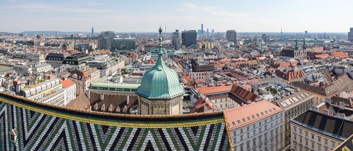 A rooftop view across a city of tiled roofs. In the foreground a steep roof covered in glazed tiles laid in green, gold, black and white zigzags; behind it an octagonal stone turret capped with a green copper dome and a slender finial; beyond, blocks of red and grey roofs running to a hazy skyline of glass towers under a pale sky.