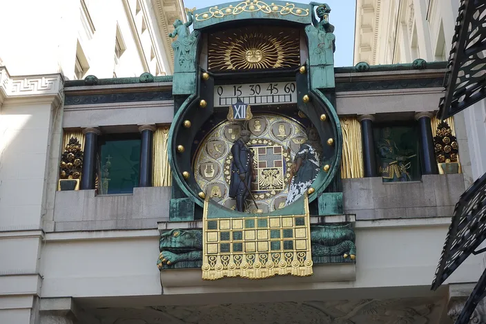 An art nouveau clock slung on a copper bridge between two buildings: a large enamelled dial ringed with gilt studs, a minute scale reading 0 15 30 45 60 above it, the Roman numeral XII marked beside the aperture, two dark bronze figures crossing the face, and a gilded openwork panel hanging beneath the whole thing.