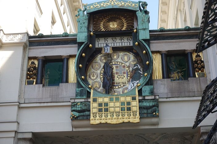 An art nouveau clock slung on a copper bridge between two buildings: a large enamelled dial ringed with gilt studs, a minute scale reading 0 15 30 45 60 above it, the Roman numeral XII marked beside the aperture, two dark bronze figures crossing the face, and a gilded openwork panel hanging beneath the whole thing.