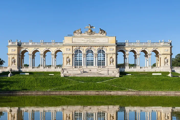 A long single-story colonnade of round arches standing on a grassy crest, a raised triumphal arch and a sculpted eagle trophy at its center, stone trophy groups on the lawn to either side, and the whole building doubled in the still water of the pool below it under a cloudless sky.