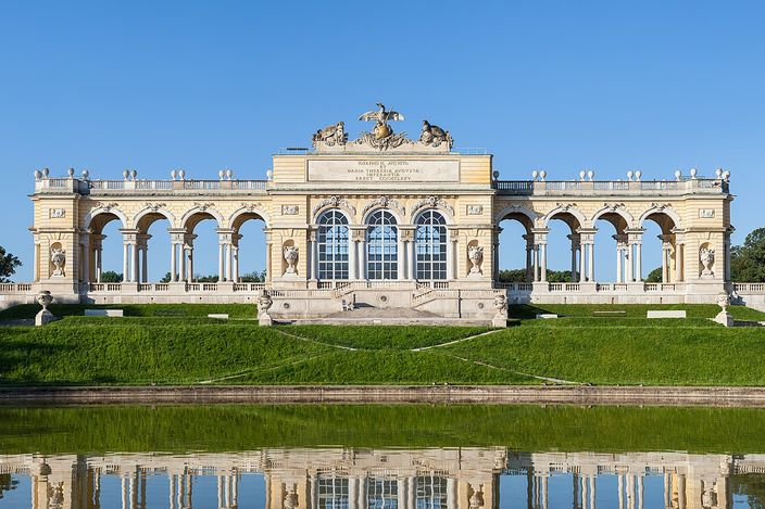 A long single-storey colonnade of round arches standing on a grassy crest, a raised triumphal arch and a sculpted eagle trophy at its centre, stone trophy groups on the lawn to either side, and the whole building doubled in the still water of the pool below it under a cloudless sky.