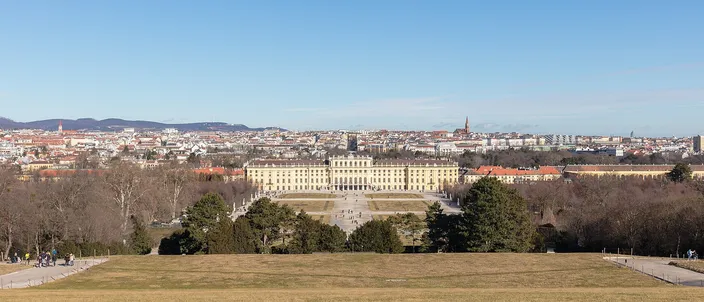 The long yellow garden front of Schoenbrunn palace seen from the hill above it, clipped hedges and a parterre running down towards the building, and behind it the whole of Vienna spread flat to a line of low wooded hills under a pale winter sky; a few walkers stand on the grass in the foreground.