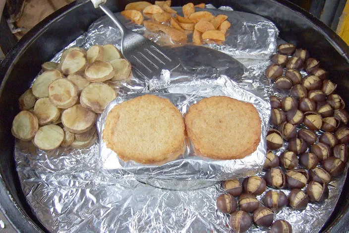 The round griddle of a Viennese street chestnut roaster seen from above: roasted chestnuts down one side, potato slices and wedges on the other, two potato pancakes in the middle