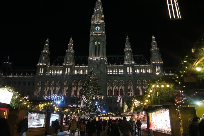Vienna's neo-Gothic City Hall lit up at night above the Christmas market on Rathausplatz, a tall decorated tree in front of it and visitors walking between the lit wooden huts
