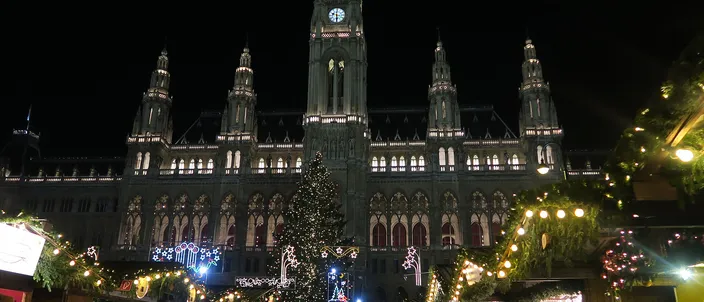 Vienna's neo-Gothic City Hall lit up at night above the Christmas market on Rathausplatz, a tall decorated tree in front of it and visitors walking between the lit wooden huts