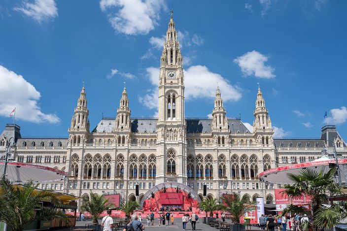 Vienna's neo-Gothic Rathaus (City Hall) with its lacy spires and central tower behind the Rathausplatz, palm trees and event stalls in the foreground under a blue sky