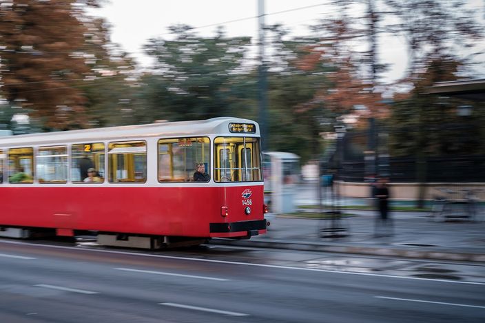A red-and-white Vienna tram on route 2 sweeping along the Ringstrasse at Dr.-Karl-Renner-Ring, caught in a panning shot so the tram is sharp and the autumn trees, railings and passers-by behind it blur into streaks; overhead wires cross the pale sky.