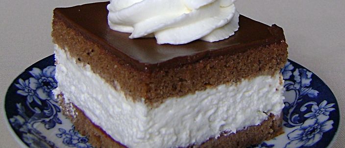 A slice of wuzetka on a blue-and-white ceramic plate: a dark cocoa sponge base, a thick layer of whipped cream, a chocolate glaze on top and a piped rosette of whipped cream.