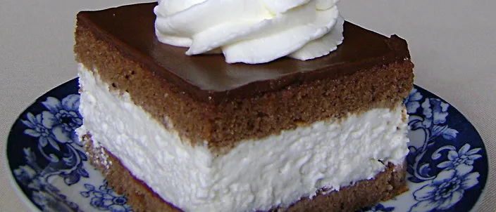 A slice of wuzetka on a blue-and-white ceramic plate: a dark cocoa sponge base, a thick layer of whipped cream, a chocolate glaze on top and a piped rosette of whipped cream.