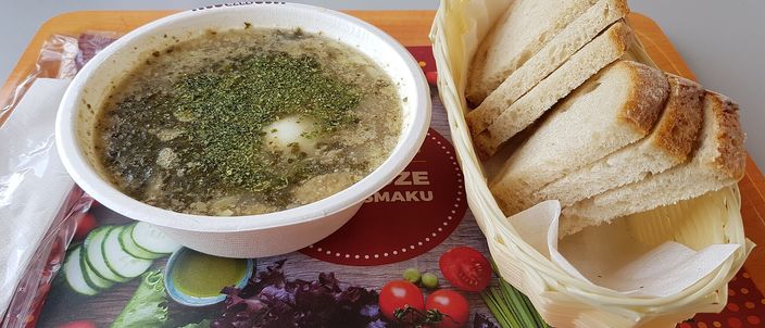 A bowl of żurek — a pale, cloudy sour rye soup flecked with green herbs and pieces of sausage — served in a plain white bowl with slices of bread alongside.