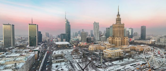 Elevated view over central Warsaw at dusk: the spired Palace of Culture and Science stands among a cluster of modern glass towers, with snow dusting the streets and pink sky behind
