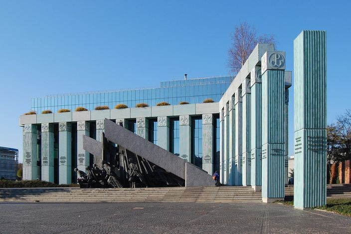 The Warsaw Uprising Monument on Krasiński Square: bronze fighters emerging from beneath a tilted slab of dark stone, with the ribbed glass and green columns of the Supreme Court building standing behind it under a clear sky