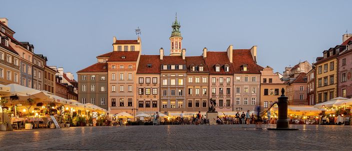 Warsaw's Old Town Market Square at dusk, its tall pastel burgher houses with red-tiled roofs closing three sides, cafe awnings lit beneath them and the bronze mermaid with her sword raised on the open cobbles