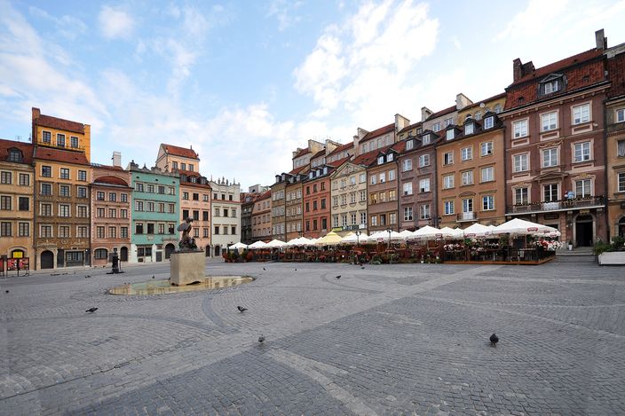 Warsaw's Old Town Market Square almost empty in early morning light, its ring of tall narrow burgher houses in ochre, red and green closing two sides, furled cafe umbrellas along the far edge, and the dark bronze mermaid on her plinth in a shallow basin on the open cobbles