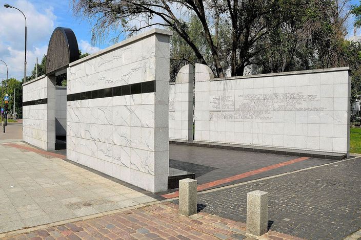 The Umschlagplatz monument in Warsaw seen from the corner: low walls of white marble banded with black, enclosing an open paved court, with a dark rounded stone canopy rising at the far end and trees in full leaf behind, granite bollards along the pavement in front