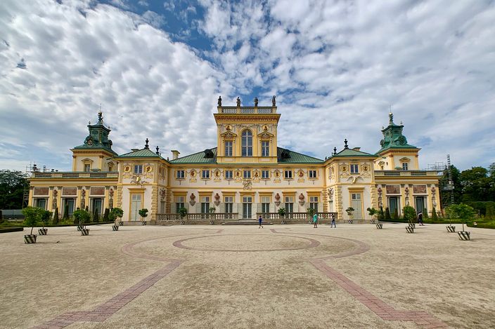 The garden facade of Wilanów Palace: a long two-storey baroque range in pale yellow with white pilasters, sculpted reliefs and a green copper roof, twin corner pavilions with cupolas, standing behind a wide gravel forecourt patterned in brick under a mackerel sky