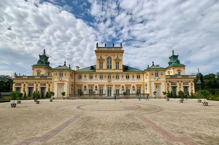 The garden facade of Wilanów Palace: a long two-story baroque range in pale yellow with white pilasters, sculpted reliefs and a green copper roof, twin corner pavilions with cupolas, standing behind a wide gravel forecourt patterned in brick under a mackerel sky