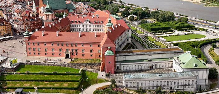 Oblique aerial view of Warsaw's Royal Castle: the long red-roofed brick range and its green-domed clock tower on the edge of the Old Town, the escarpment falling away in terraced lawns to a road and the Vistula beyond, with the Old Town's tiled roofs and Castle Square crowded with people at the left