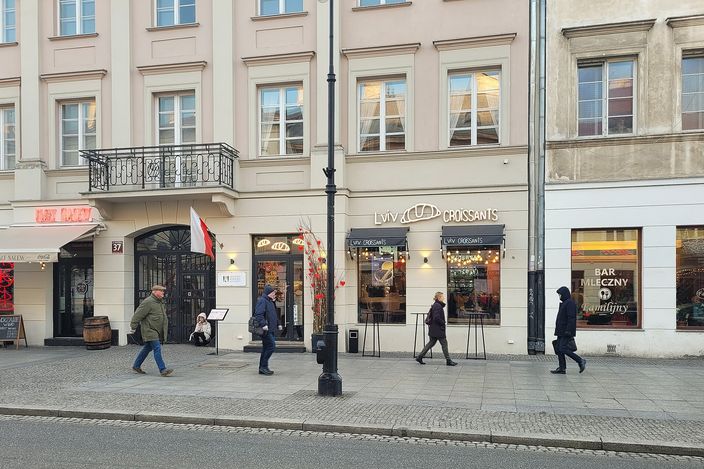 Pedestrians walking past a milk bar (Bar Mleczny) and a Lviv Croissants café at street level in nineteenth-century townhouses on Nowy Świat, Warsaw, on a clear winter day.