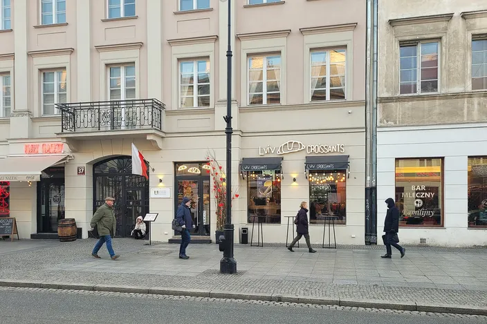 Pedestrians walking past a milk bar (Bar Mleczny) and a Lviv Croissants café at street level in nineteenth-century townhouses on Nowy Świat, Warsaw, on a clear winter day.