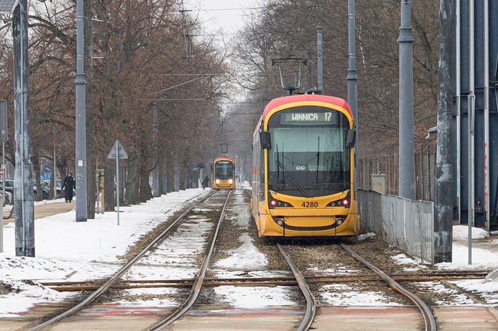 Two red-and-yellow Warsaw trams passing on twin tracks down a tree-lined avenue with light snow on the ground; the near tram, numbered 4280, shows route 17 to Winnica.