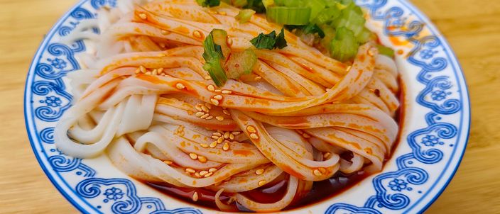 A bowl of Xi'an liangpi — wide translucent rice-noodle ribbons coated in glossy red chili oil, topped with chopped scallion and sesame seeds, in a blue-and-white porcelain bowl on a wooden table.