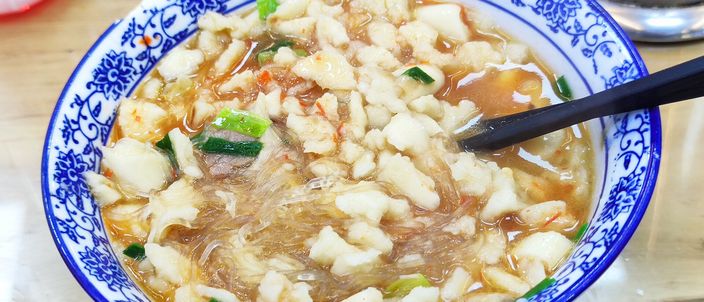 A blue-and-white porcelain bowl of yangrou paomo filled with grain-sized crumbled flatbread, glass noodles, scallions and a dollop of red chilli paste, black chopsticks resting across the rim.