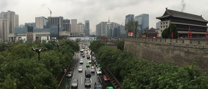 A grey stone city-wall rampart and traditional watchtower on the right, with Xi'an's modern high-rise skyline and a busy tree-lined boulevard below on the left, photographed from atop the wall on an overcast day.