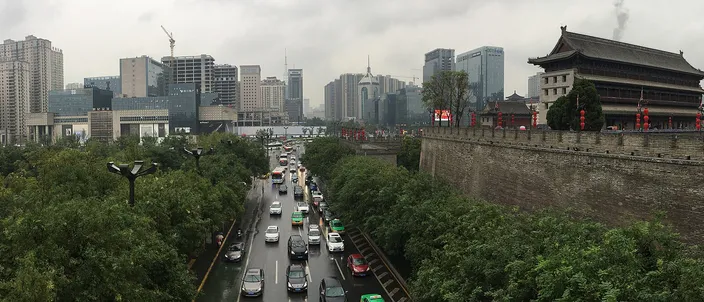 A gray stone city-wall rampart and traditional watchtower on the right, with Xi'an's modern high-rise skyline and a busy tree-lined boulevard below on the left, photographed from atop the wall on an overcast day.