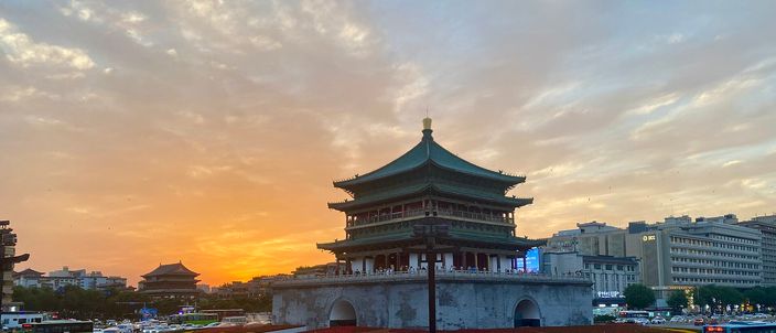 Xi'an's Bell Tower silhouetted against an orange sunset sky, ringed by a busy traffic circle and the city's buildings.