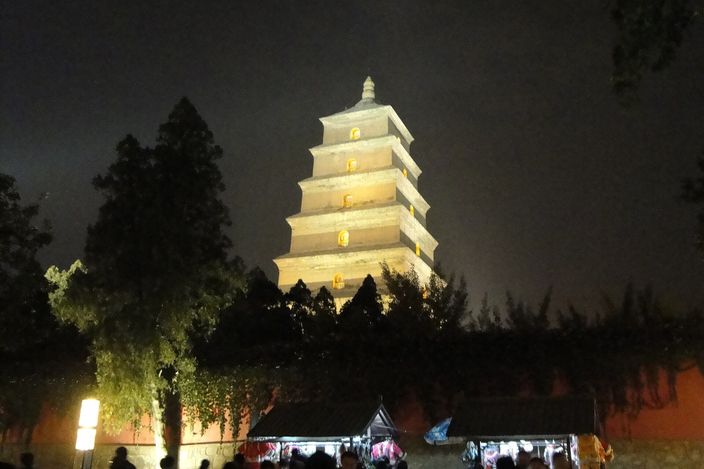 The Big Wild Goose Pagoda illuminated at night, its seven brick storeys lit gold above a lantern-lit market street and evening crowd at its base.