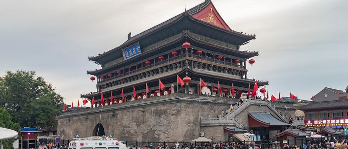 The Drum Tower of Xi'an on an overcast day, its triple-eaved timber roof hung with red lanterns and flags, crowds and a police van in the square below.
