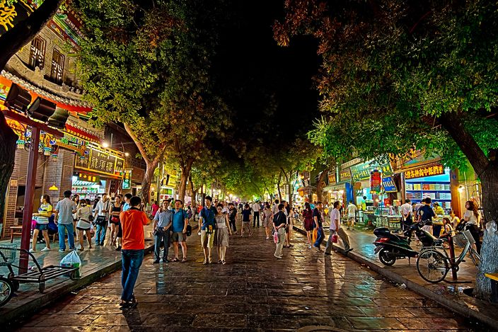 A crowded pedestrian lane in Xi'an's Muslim Quarter at night, strings of red lanterns and mosque-style tiled eaves overhead, food stalls and shopfronts lit up on both sides, people walking down the middle of the street.