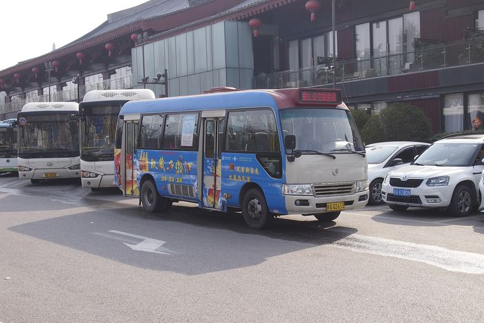 A blue-and-white city bus on the Qujiang route parked outside a red Tang-style building decorated with lanterns, other buses and parked cars alongside.