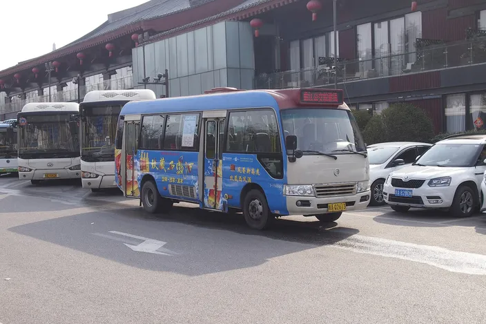 A blue-and-white city bus on the Qujiang route parked outside a red Tang-style building decorated with lanterns, other buses and parked cars alongside.