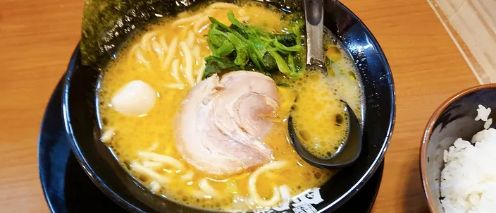 A bowl of iekei ramen: thick straight noodles in an opaque tan pork-and-soy broth, topped with a slice of chashu pork, boiled spinach and sheets of nori standing against the rim, with a spoon in a dark bowl on a wooden counter.