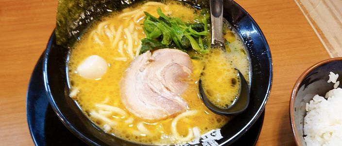 A bowl of iekei ramen: thick straight noodles in an opaque tan pork-and-soy broth, topped with a slice of chashu pork, boiled spinach and sheets of nori standing against the rim, with a spoon in a dark bowl on a wooden counter.