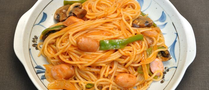 A plate of Napolitan: glossy ketchup-red spaghetti pan-fried with strips of green pepper and pieces of sausage, served on a patterned plate.