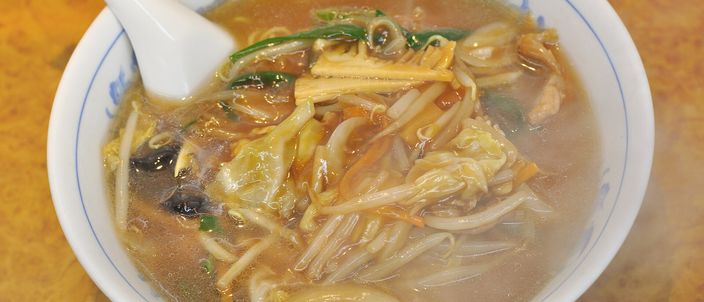 A bowl of sanmamen: soy-coloured noodle soup topped with a starch-thickened tangle of bean sprouts, cabbage, wood-ear mushroom and thin pork, with a white spoon resting in a Chinese-patterned bowl.