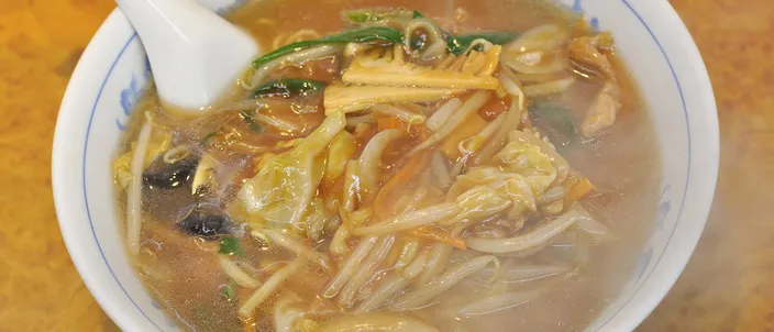 A bowl of sanmamen: soy-colored noodle soup topped with a starch-thickened tangle of bean sprouts, cabbage, wood-ear mushroom and thin pork, with a white spoon resting in a Chinese-patterned bowl.