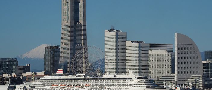 The Yokohama Minato Mirai waterfront skyline in daylight — the tapered Landmark Tower, the Cosmo Clock Ferris wheel and the curved Queen's Square towers, with a white cruise ship berthed in front and the snow-capped cone of Mount Fuji faint to the left.