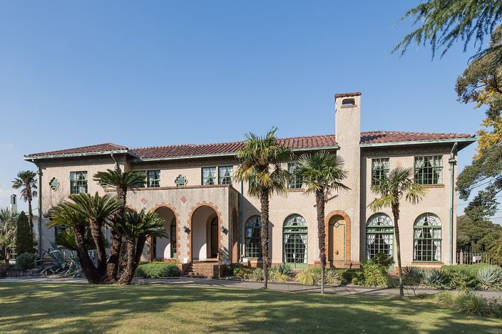 A two-storey cream stucco house with an orange pantile roof, a tall chimney and an arched ground-floor porch, standing behind palms and a mown lawn under a clear blue sky.