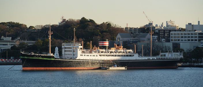 The black-hulled 1930 liner Hikawa Maru moored at a quay in low evening sun, her white superstructure and buff funnels set against a wooded hillside and low city buildings, with harbour water across the foreground.